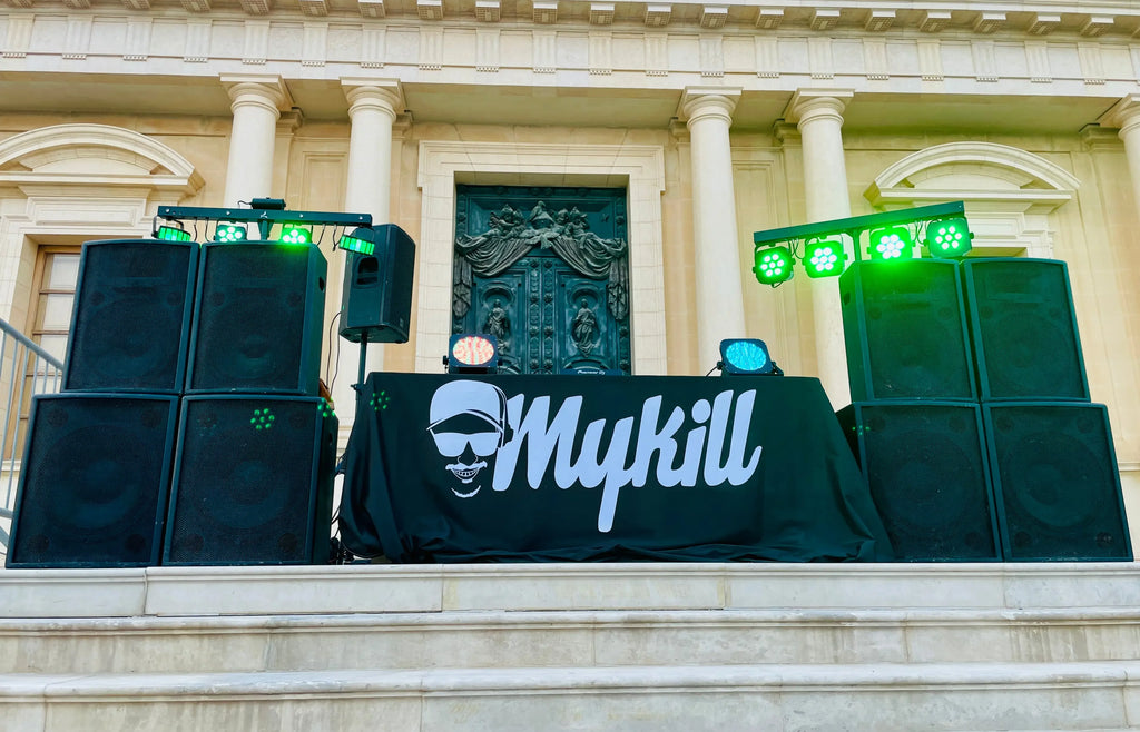 DJ setup with speakers and green lights at a grand building, featuring Mykill branding for event entertainment.