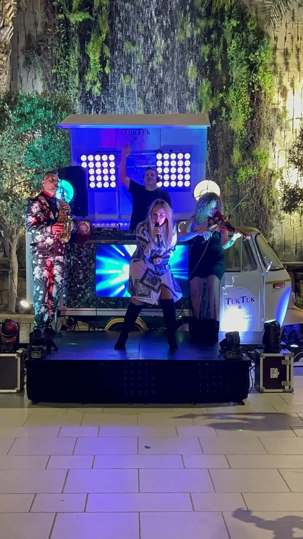 Live band with singer, saxophonist, violinist, and DJ performing on stage at night with TukTuk backdrop